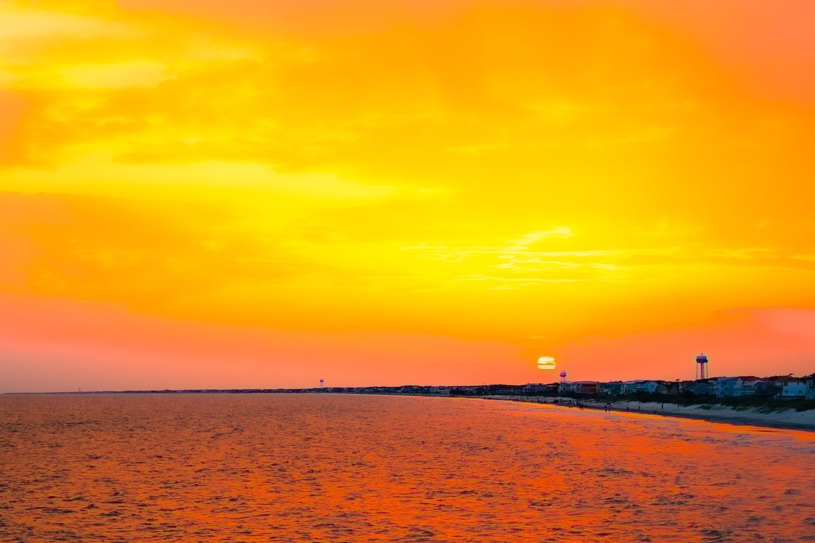 From the Oak Island Pier, the sun sinks like a molten ember into a sea of orange and gold, washing the shoreline in warm, electric color. The curve of the beach, dotted with water towers and tiny silhouettes of evening walkers, feels like the perfect front‑row seat to one of Oak Island’s signature sunsets.