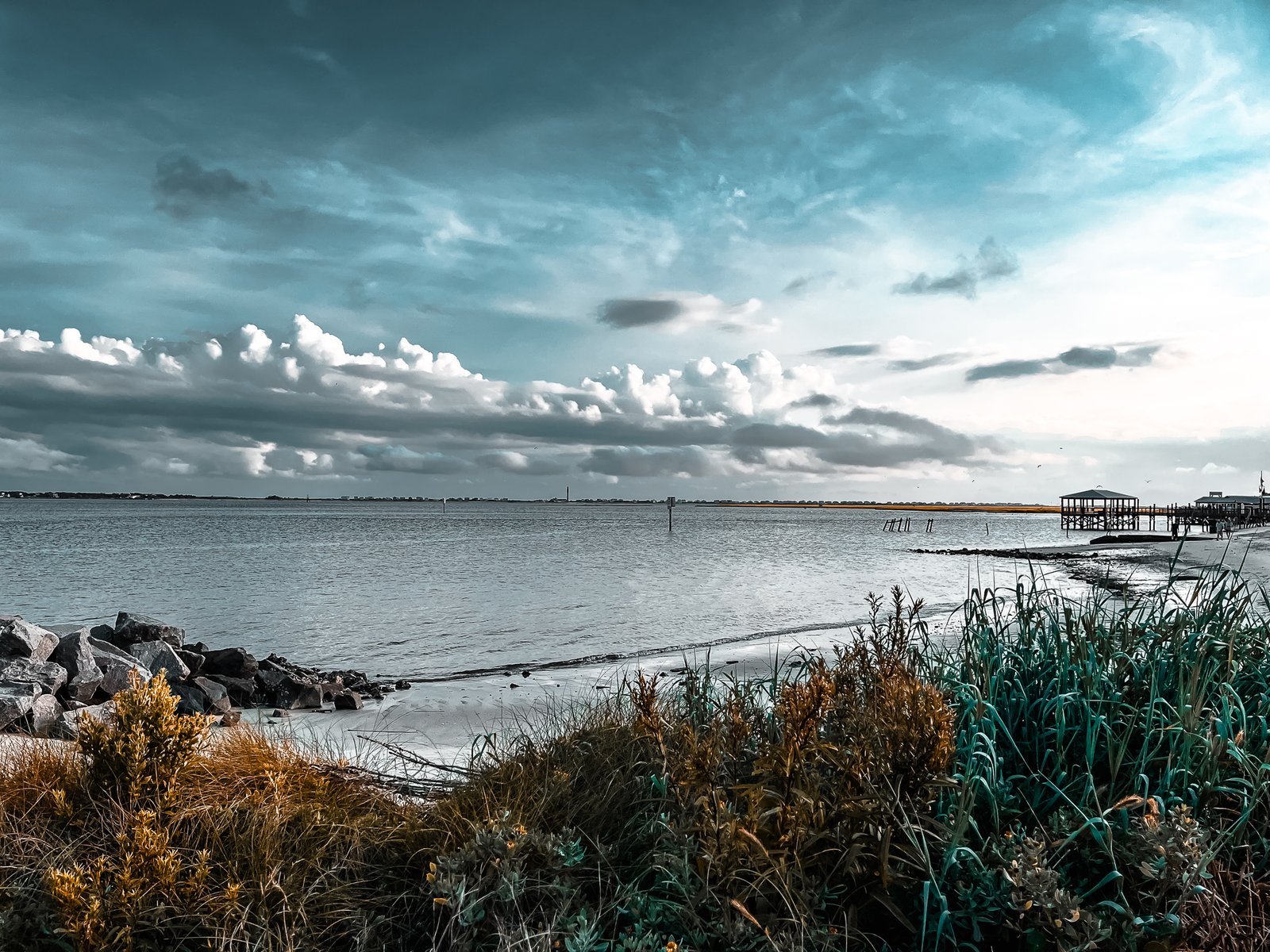 This artistic interpretation of Southport’s waterfront captures the moody beauty of the Cape Fear River on a day of dramatic skies . Shot from the 9-acre Waterfront Park, the image looks across the river toward Oak Island’s eastern tip, where historic Fort Caswell stands guard at the mouth of the Cape Fear River . The iconic Oak Island Lighthouse, visible from Southport’s waterfront, rises on the horizon where the river meets the Atlantic Ocean .
Selective color processing brings golden warmth to the native coastal vegetation in the foreground while rendering the water, sky, and fishing piers in monochromatic tones—creating a dreamlike quality that bridges memory and reality . The composition tells the story of centuries of maritime history, where Fort Caswell has protected this strategic waterway since 1838, guarding the approach to Wilmington 20 miles upriver .
Rocky jetties anchor the left side of the frame, protecting the shoreline where the Cape Fear River widens into its estuary. Wooden piers extend into the distance, inviting anglers to test the deep waters that flow past Fort Caswell’s eastern promontory . The artistic treatment emphasizes the eternal nature of this coastal landscape—the ever-changing sky above constant waters, wild grasses persisting season after season, and the timeless rhythm of river life.
