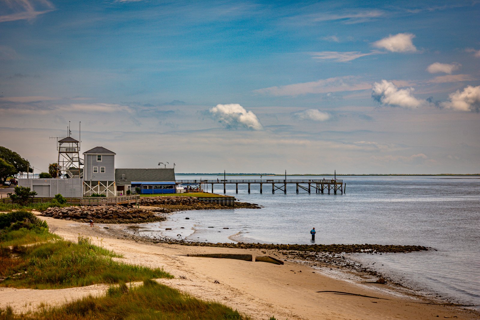 A lone angler wades in the shallow waters along Southport’s Cape Fear River shoreline, casting into the productive tidal zone while the Coast Guard’s distinctive observation tower stands watch in the background . This scene captures the peaceful coexistence of recreation and maritime safety that defines Southport’s waterfront character . The historic U.S. Coast Guard Station Oak Island, originally established as a Life-Saving Service station in 1891, continues its mission of protecting mariners navigating these strategic waters where the Cape Fear River meets the Atlantic .
The Southport City Pier extends into the river, a beloved spot for anglers and sightseers enjoying the tranquil views across to Oak Island . Rocky jetties protect the sandy shoreline where beach grasses transition to the wide expanse of water. The composition tells a quiet story of coastal Carolina—where everyday life unfolds against a backdrop of maritime history, where the Coast Guard’s six waterborne assets stand ready just beyond the channel, and where a fisherman can still find solitude at low tide .
Bright blue skies with scattered clouds paint the scene in natural coastal light, while the calm river reflects the peaceful rhythm of this charming waterfront town 